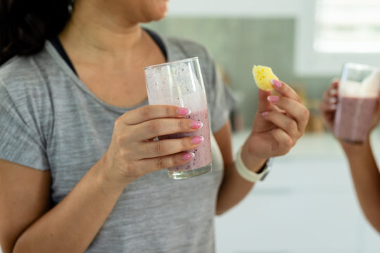 Diverse women in gray top sharing berry smoothies in kitchen, holding pineapple chunk and tracker