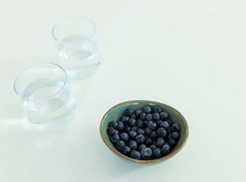 Small ceramic bowl is holding blueberries on pale tabletop near two water glasses, copy space