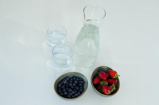 Glass carafe sitting partly filled on white table, glasses beside bowls of blueberry and strawberry