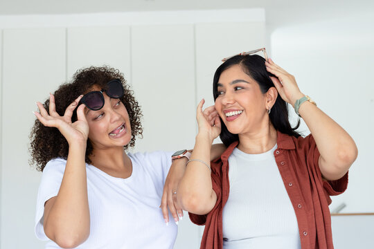 Diverse female friends standing smiling and trying on dark sunglasses showing watches in kitchen