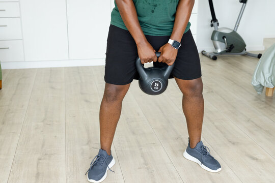 Adult African man holding 5 kg kettlebell between legs in home workout corner, wearing smartwatch