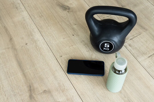 Black kettlebell sitting on light wood plank floor with smartphone and water bottle nearby