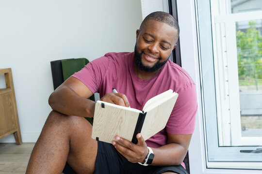 African American man sitting on floor at home writing in woodgrain notebook holding pen