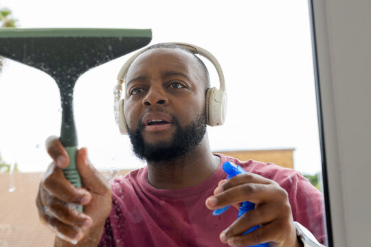 Man cleaning window outside wearing maroon T-shirt, headphones, holding squeegee and spray bottle