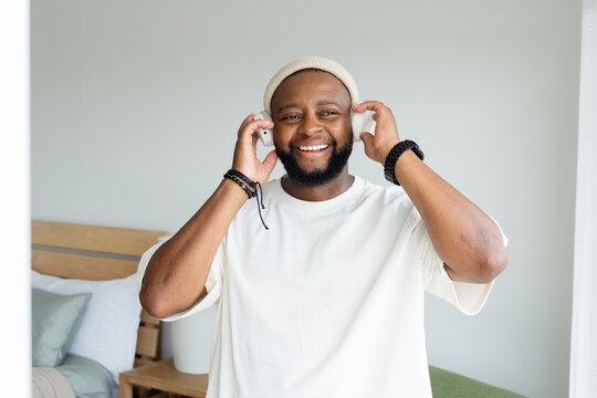 Man smiling and holding headphones while listening with smartphone in bright bedroom wearing beanie