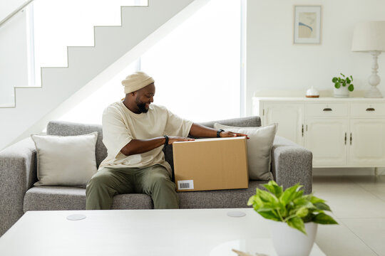 African American man sitting on gray sofa smiling while holding cardboard shipping box