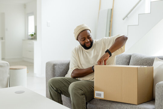 African American man wearing beanie sitting on gray sofa in living room opening cardboard box