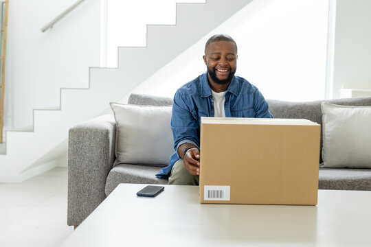 Adult African American man sitting in living room wearing denim jacket opening parcel near phone