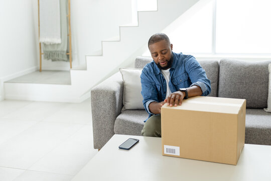 Man sitting on gray sofa unpacking cardboard shipping box while checking smartphone in living room