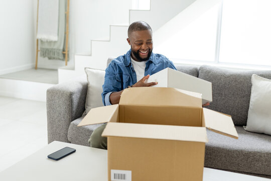 African man in denim smiling lifting white box on grey sofa near shipping box and smartphone