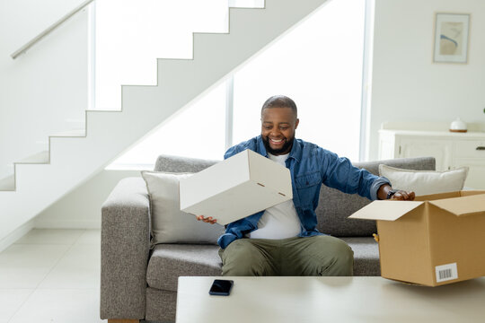 African American man sitting at home on sofa holding white box beside shipping box with smartphone