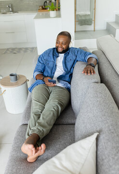 Adult African male reclining on sofa at home, wearing denim jacket and headphones with smartphone