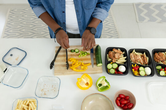 Adult African man in denim shirt chopping and arranging vegetables at counter with knife, trays