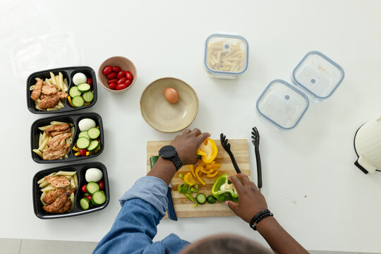 African American man slicing bell peppers on cutting board, prepping meal-prep containers with eggs