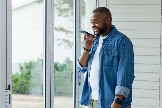 Adult African man in denim shirt holding smartphone near sliding glass door at home, copy space