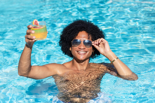 African man smiling, holding cocktail and adjusting aviator sunglasses, shirtless in sunny pool