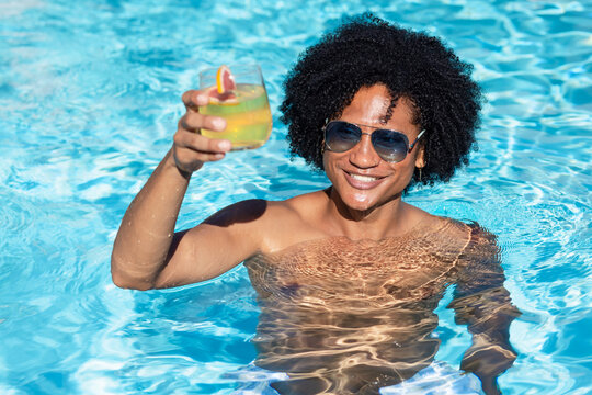 African adult male smiling, shirtless wearing aviators, holding cocktail with citrus-wedge in pool