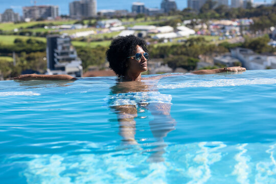 Man leaning in rooftop infinity pool wearing mirrored sunglasses bracelet swimwear viewing skyline