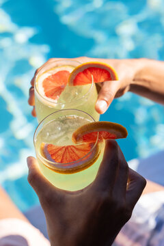 Two hands are holding short cocktail glasses over pool, showing red citrus wheels and ice