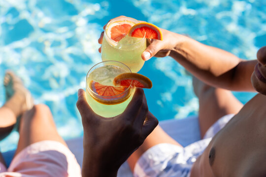 Two men are clinking cocktails and sitting on pool edge with sparkling blue water