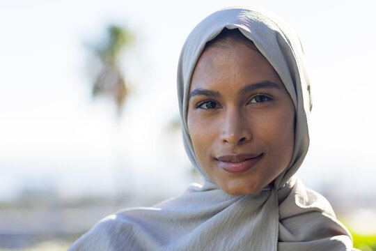 Woman wearing light gray headscarf and draped garment posing outdoors with blurred palm tree