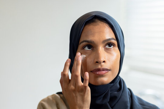 African American woman smoothing eye cream with fingertip in room near window blinds wearing hijab