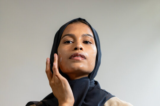 African American female wearing hijab, touching cheek with cream and fingertips against gray wall