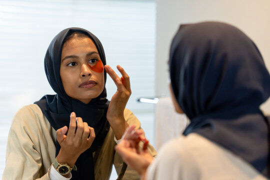 Woman wearing hijab and tan top applying orange eye patch holding jar at bathroom mirror