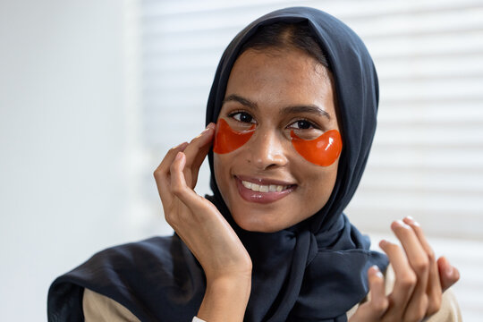 Woman wearing dark navy headscarf applying orange under-eye hydrogel patches by window blinds