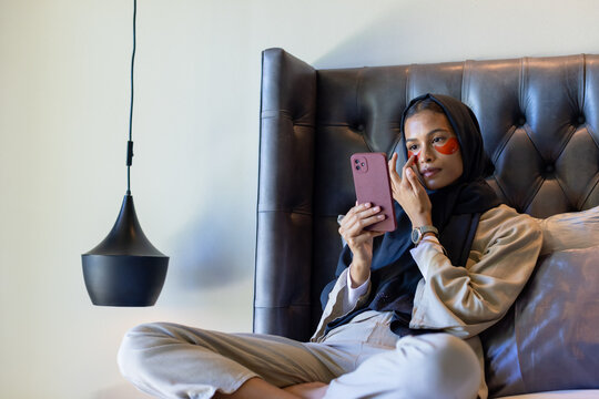 Adult woman leaning on tufted headboard in bedroom holding pink phone touching orange eye patches