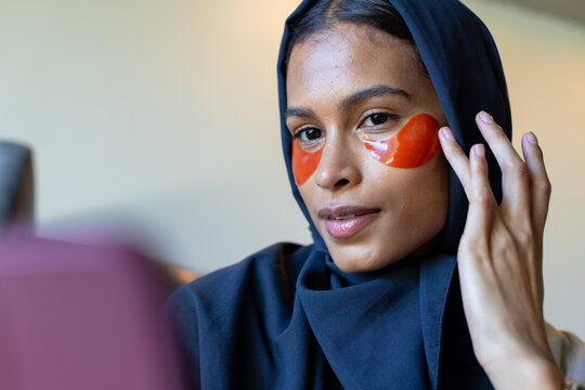 Woman wearing dark hijab touching orange gel under-eye patches sitting at vanity with pink device