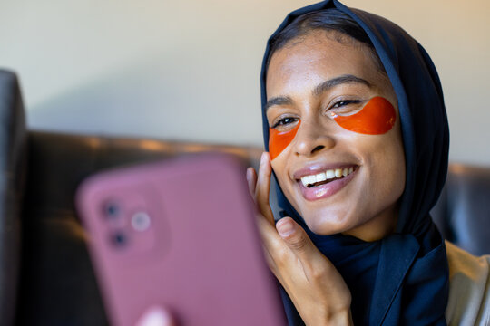 Smiling woman wearing dark scarf and orange eye patches sitting on leather sofa holding pink phone