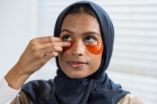 African-American woman in navy hijab adjusting orange hydrogel patch under eye at vanity