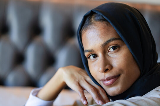 Adult African woman wearing dark hijab resting chin on hand, sitting on tufted sofa, copy space