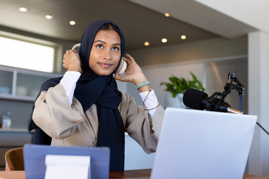 Female in dark headscarf adjusting over-ear headphones in home studio with laptop microphone