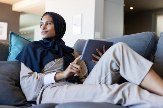 Woman reclining on sofa with teal pillow holding dark tablet tapping screen wearing navy headscarf