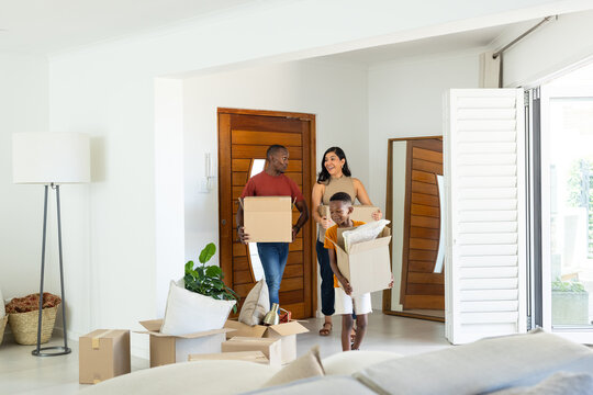 Diverse family carrying cardboard boxes and pillow while moving into bright living area