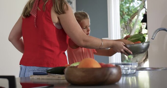 Woman and child girl moving colander rinsing greens under faucet at kitchen sink making meal