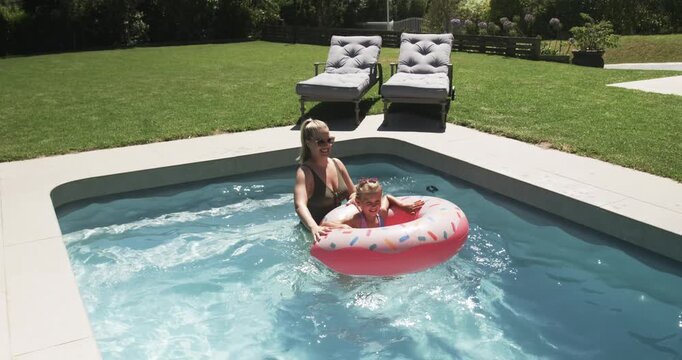 Adult female guiding child in swimsuits at pool gently pushing and steadying pink donut float