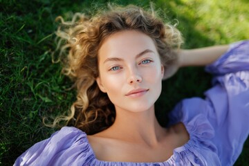 Naklejka premium Young caucasian female adult portrait lying in grass wearing lavender dress with curly hair blue eyes natural makeup soft sunlight outdoor relaxed mood