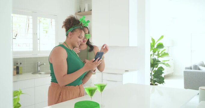Diverse women pals posing selfies in kitchen, phone held up placing green party props and drinks