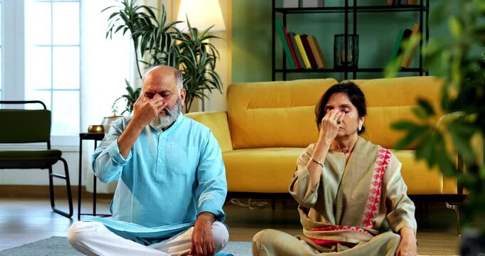 Indian senior couple doing anulom vilom pranayama or yoga at home in the morning sitting with folded legs in modern living room, practicing mindful breathing exercise for health and fitness