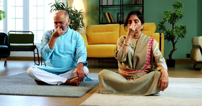 Indian senior couple doing anulom vilom pranayama or yoga at home in the morning sitting with folded legs in modern living room, practicing mindful breathing exercise for health and fitness