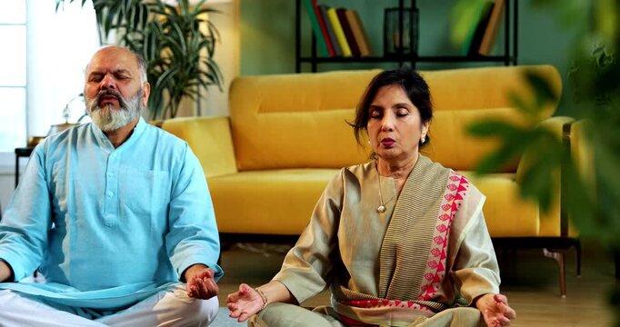 Indian senior couple doing omkar pranayama or yoga at home in the morning sitting with folded legs in modern living room chanting aum, practicing mindful breathing exercise for health