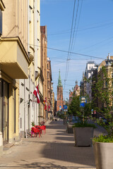 A street with a red and white building and a green spire in Riga Latvia. The street is lined with trees and has a few people walking around