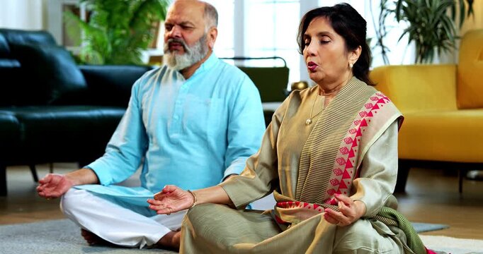 Indian senior couple doing omkar pranayama or yoga at home in the morning sitting with folded legs in modern living room chanting aum, practicing mindful breathing exercise for health