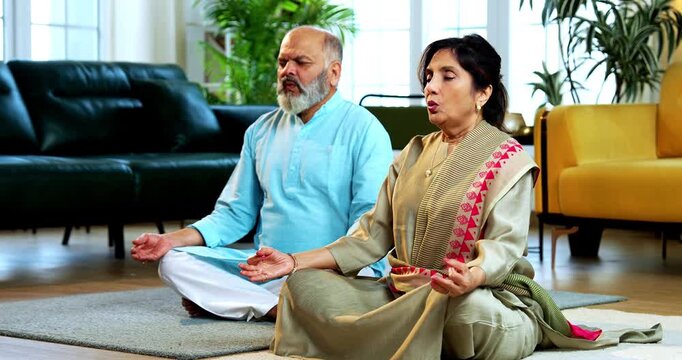 Indian senior couple doing omkar pranayama or yoga at home in the morning sitting with folded legs in modern living room chanting aum, practicing mindful breathing exercise for health