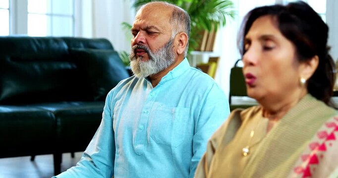 Indian senior couple doing omkar pranayama or yoga at home in the morning sitting with folded legs in modern living room chanting aum, practicing mindful breathing exercise for health
