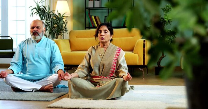 Indian senior couple doing omkar pranayama or yoga at home in the morning sitting with folded legs in modern living room chanting aum, practicing mindful breathing exercise for health
