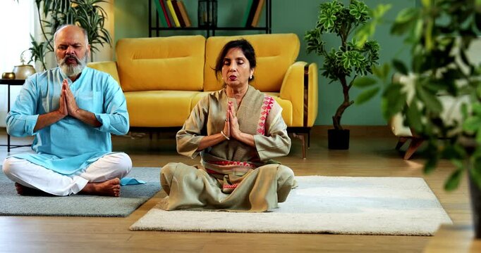 Indian senior couple doing omkar pranayama or yoga at home in the morning sitting with folded legs in modern living room chanting aum, practicing mindful breathing exercise for health